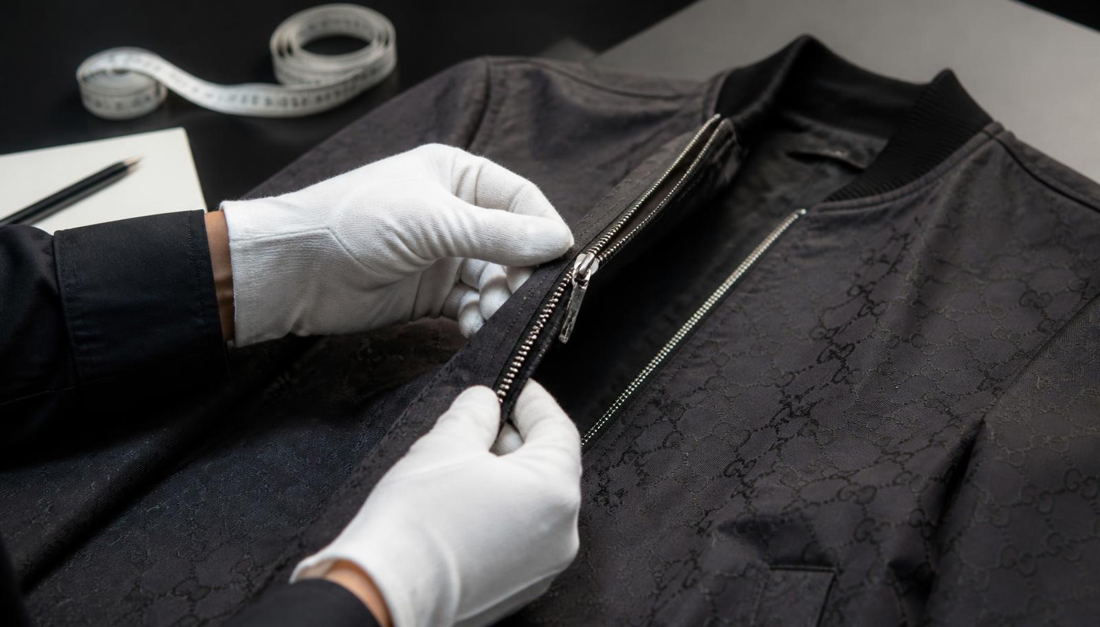 Gloved hands inspecting the zip and stitching of a dark jacket on an inspection table